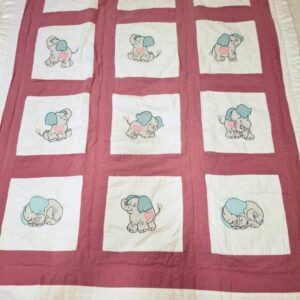 A rectangular baby crib quilt featuring a 3x4 grid of white fabric squares. Each square contains an embroidered cartoon baby elephant with teal ears and a pink heart on its side. The squares are separated by dusty rose sashing, and the entire quilt is finished with a white ruffled fabric border.