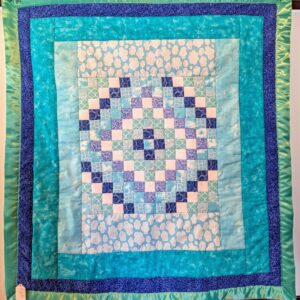 A square handmade quilt featuring a central diamond mosaic pattern made of small blue, purple, and white squares. The center is framed by light blue fabric with white cloud patterns and multiple borders in various shades of aqua, turquoise, and navy blue. The entire quilt is finished with a wide, shiny seafoam green satin border.