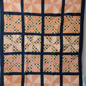 A handmade pinwheel lap quilt featuring twenty square blocks arranged in a 4x5 grid, separated by thick black sashing. The blocks alternate between solid peach-toned pinwheels and pinwheels featuring a vibrant orange, black, and white gingham plaid. The quilt is finished with a solid black border and features intricate, circular decorative top-stitching across the center of each block.