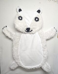 A handcrafted polar bear baby tummy time mat laid flat on a white surface, showing a smooth white fabric face and belly contrasted with a plush, curly white faux-fur textured trim on the head, arms, and legs. The bear features hand-stitched black eyes with white pupils, a black nose and mouth, and small black felt claws on its extended paws.